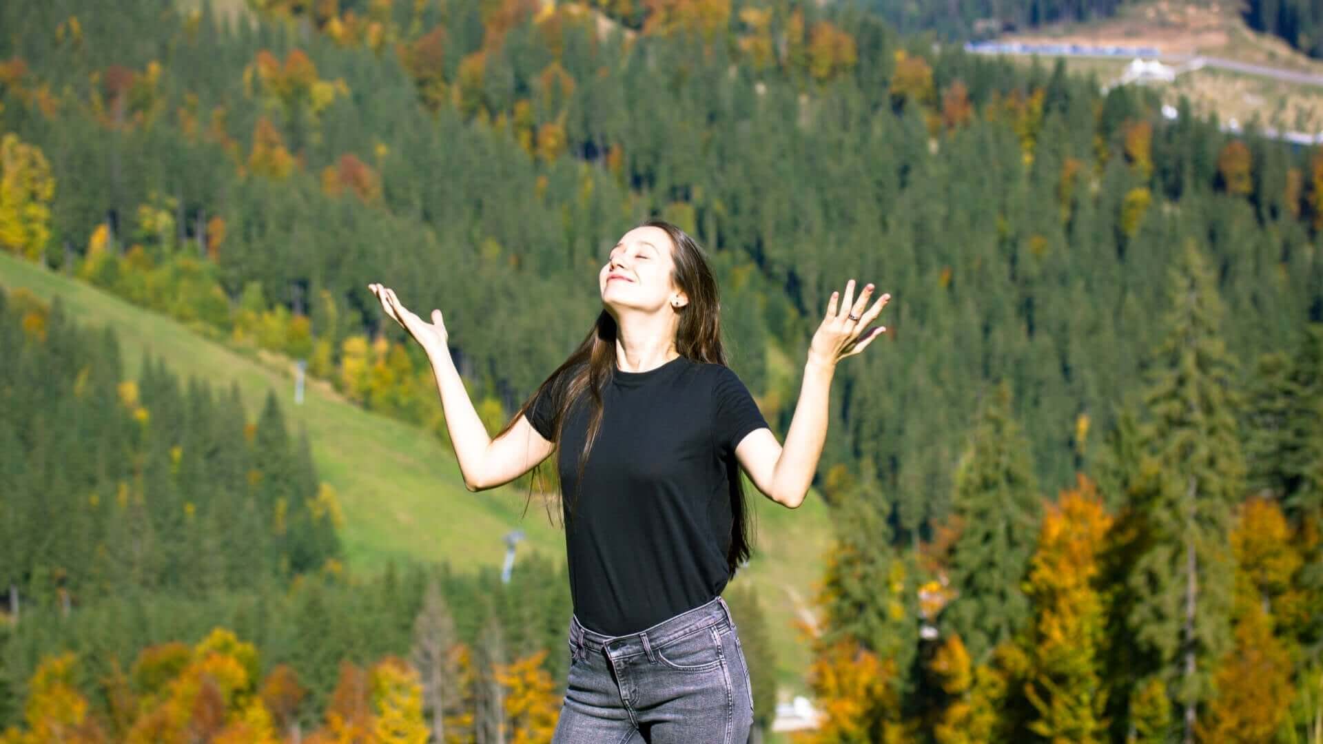 Woman in black t-shirt and gray jeans arms outstretched amid autumn trees fragrance mountain backdrop