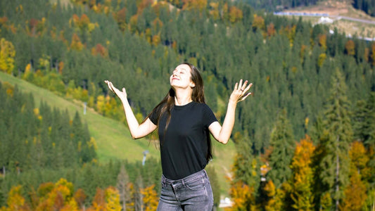 Woman in black t-shirt and gray jeans arms outstretched amid autumn trees fragrance mountain backdrop