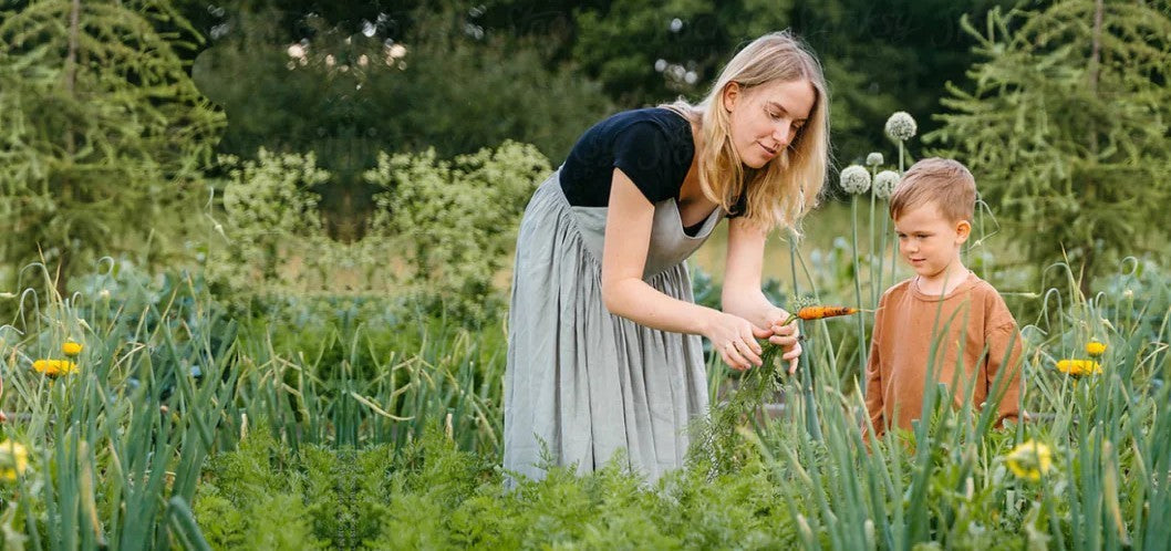 A woman in a black top and light gray skirt bends over to tend to plants in a lush green garden.